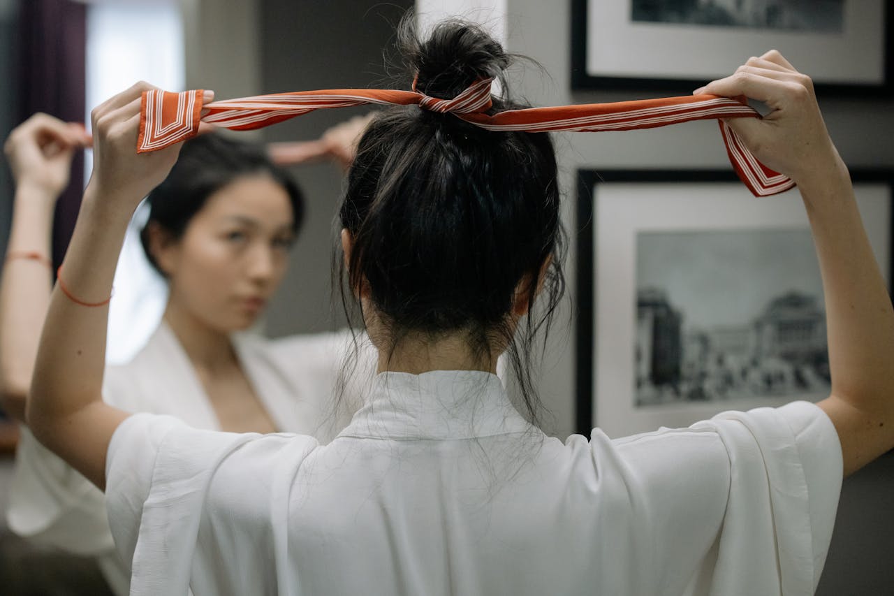 A woman in a white robe styles her hair with a red ribbon in front of a mirror indoors.